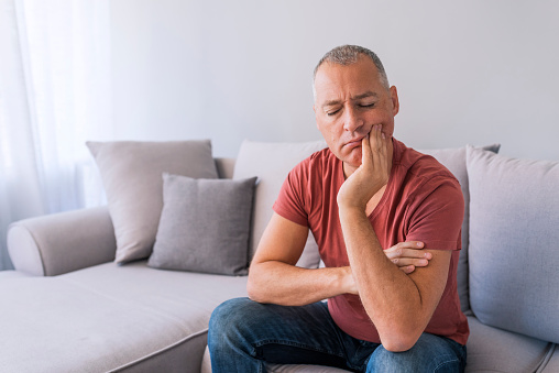 man sitting on couch with a painful expression, with eyes closed, his hand on his cheek