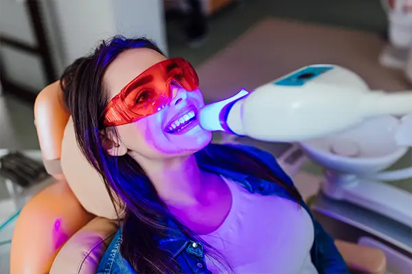 A woman in dental chair smiling while receiving teeth whitening treatment at a dental clinic.